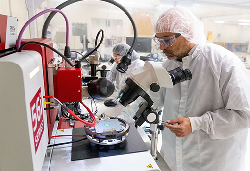 A researcher wearing a lab coat, hair protection, and eye protection works at a machine inside the Protofab lab, while another researcher works in the background.